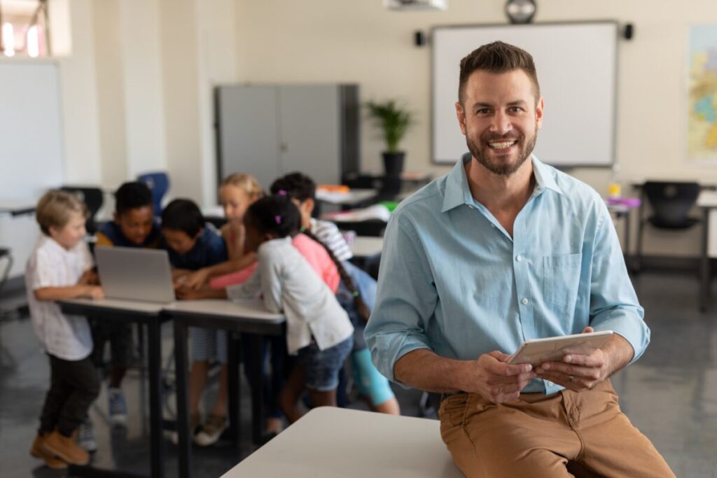 Ein Lehrer sitzt mit seinem Tablett im Klassenraum, hinter ihm arbeiten seine Schüler.