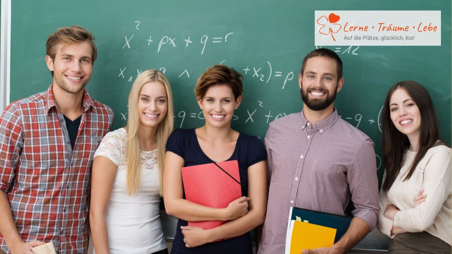 Junge Lehramtsstudierende stehen lächelnd vor einer Tafel in der Schule.